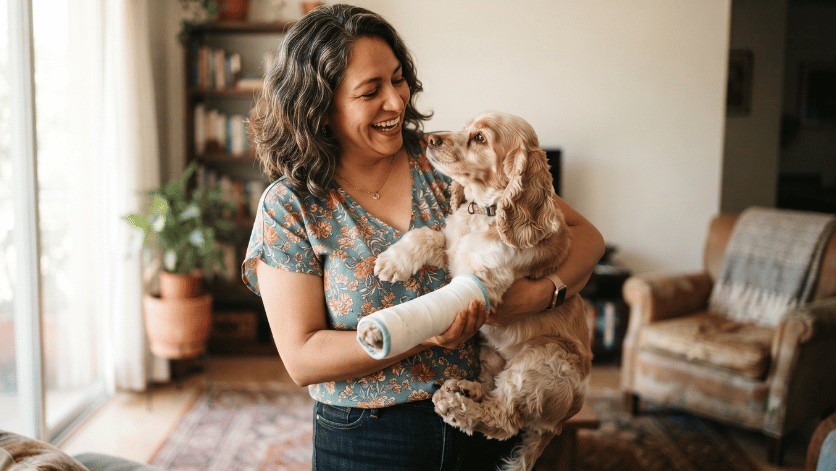 Mujer sonriendo al sostener a su perro con una patita enyesada en casa, reflejando la tranquilidad de contar con un seguro de gastos médicos veterinarios.